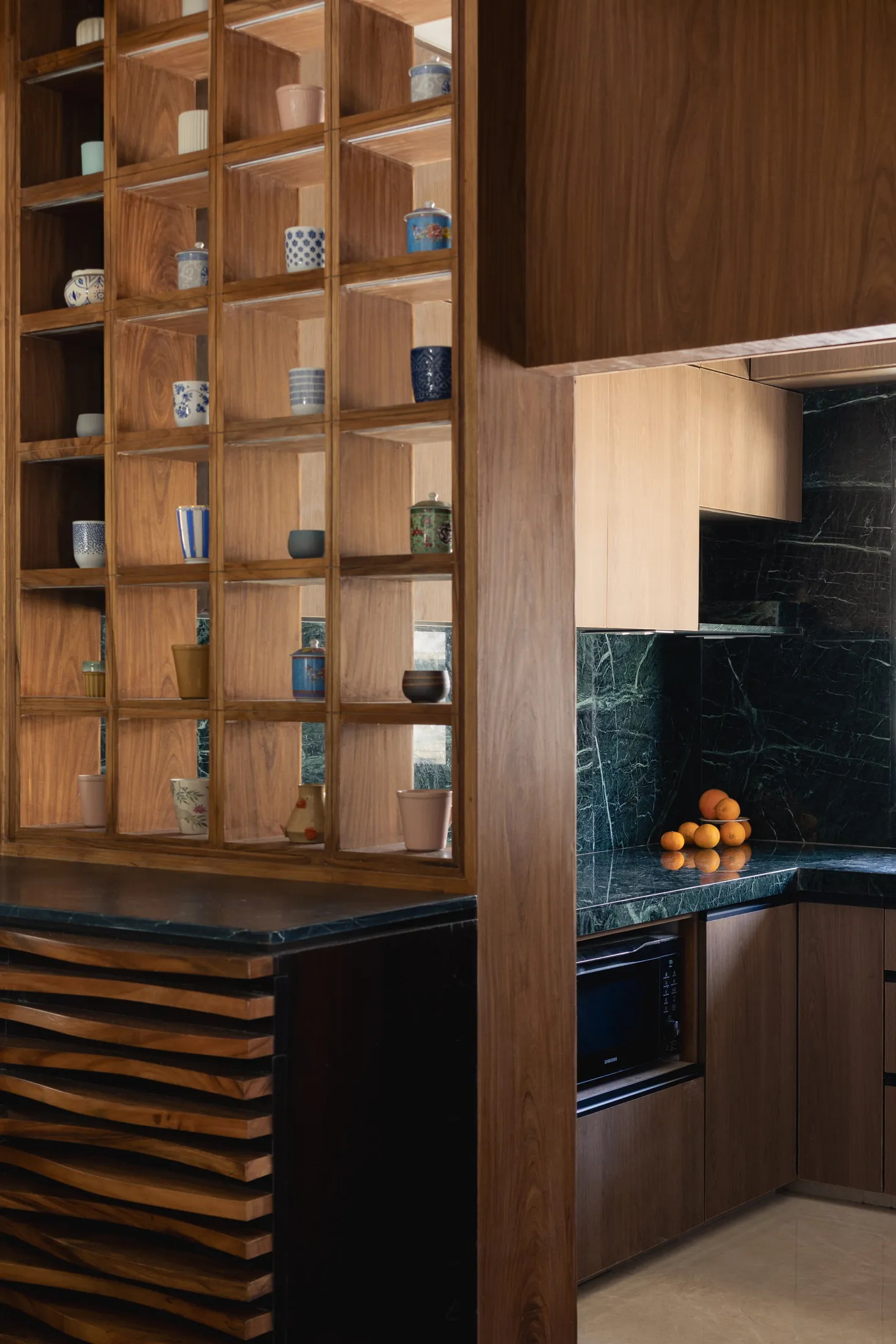 Teak display wall of open cubbies holding travel mugs and handmade pottery beside the green stone kitchen in Plum House