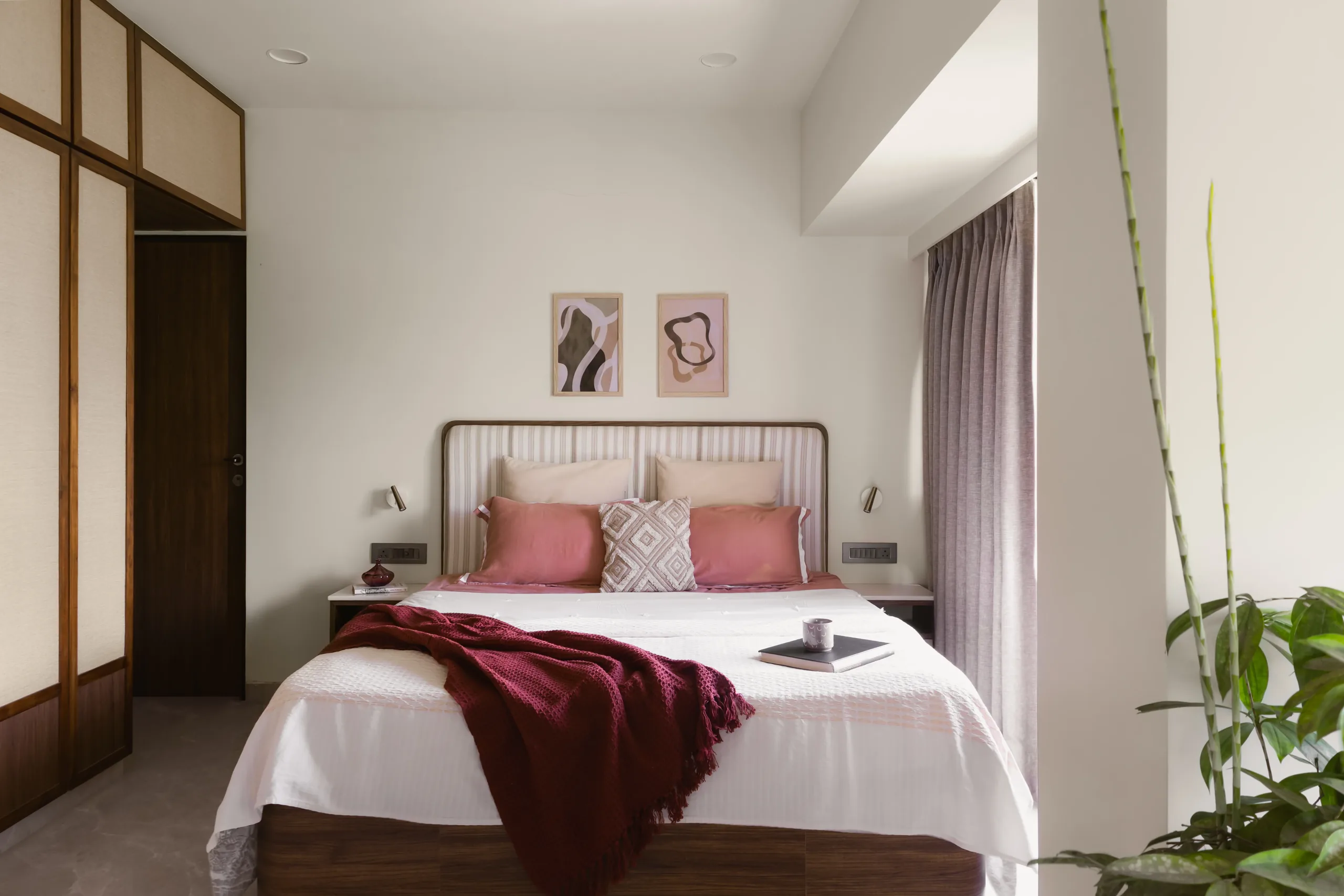 The Nest bedroom with a slim iron bed, pink cushions, burgundy throw and two framed prints