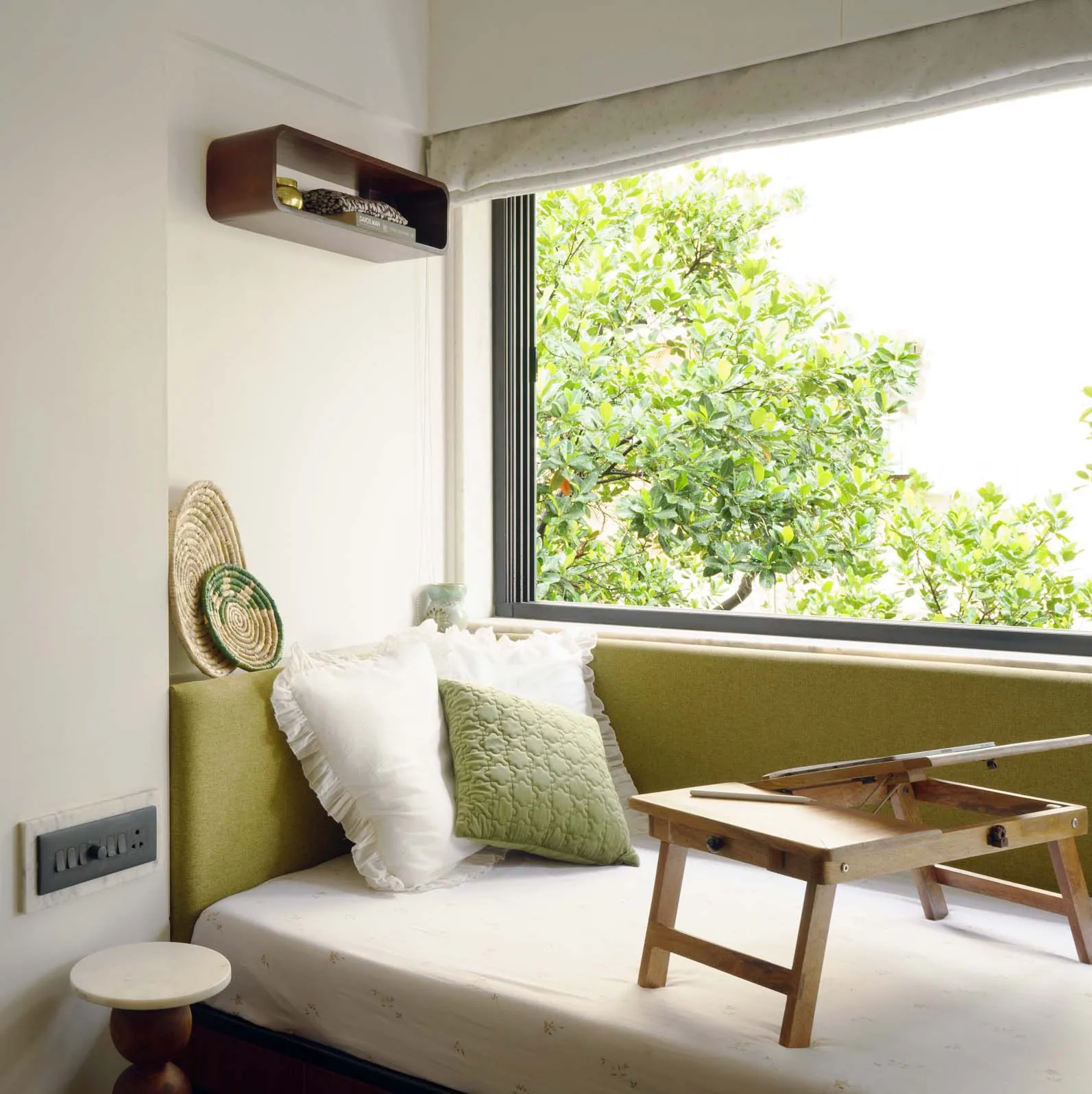 The Seed daybed with a green bolster and a folding teak tray table under a large window